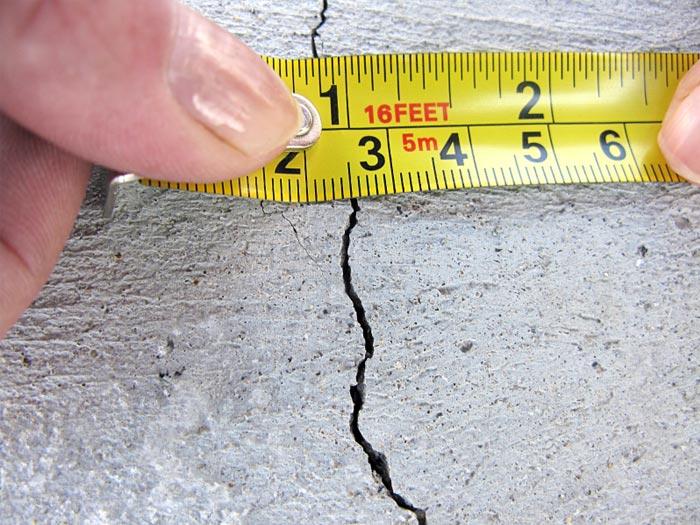 Hands Holding A Measuring Tape To Measure A Crack In A Concrete Foundation Wall, Illustrating Basement Inspection For